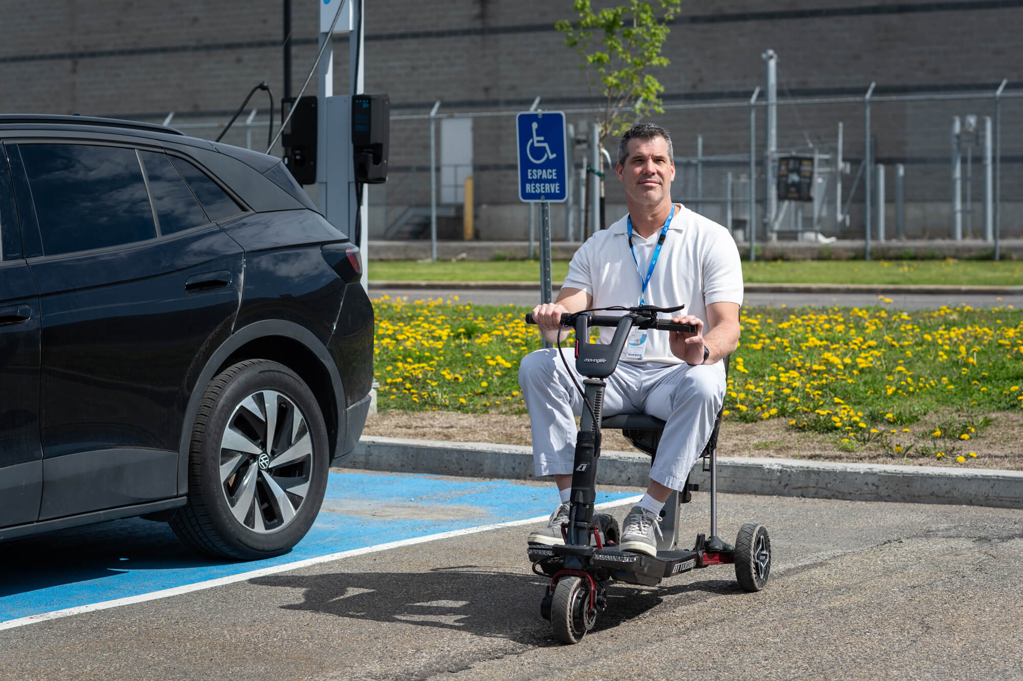 Un homme aux cheveux noirs courts, vêtu d'un polo blanc et d'un pantalon gris, est assis sur un scooter de mobilité noir sur un parking à côté d'une voiture noire.