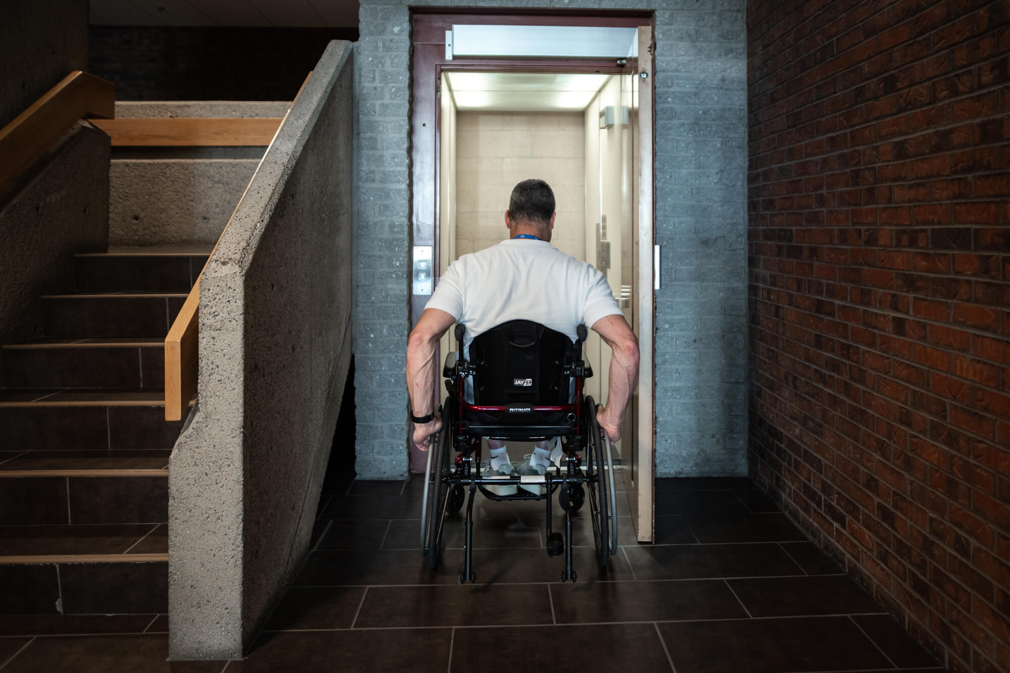 Un homme en fauteuil roulant entre dans un ascenseur dans le couloir d'un immeuble, à côté d'une cage d'escalier.