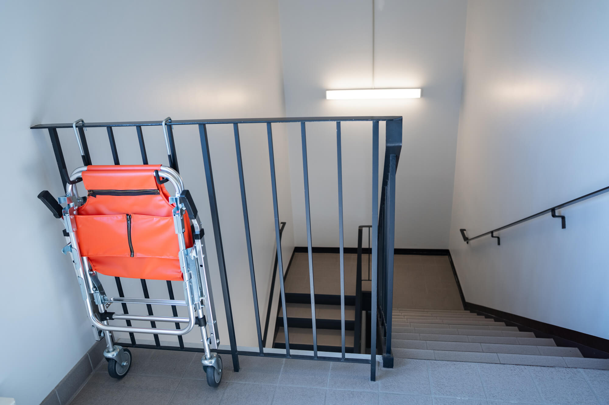 Un fauteuil roulant pliant orange repose contre une rampe en métal noir en haut d'un escalier dans le couloir d'un immeuble.