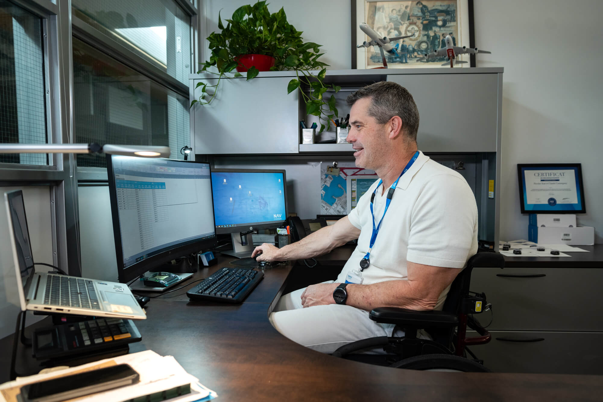 Un homme en fauteuil roulant travaille sur un ordinateur dans un bureau bien organisé, avec deux écrans, une décoration sur le thème de l'aviation et un certificat affiché sur le mur.