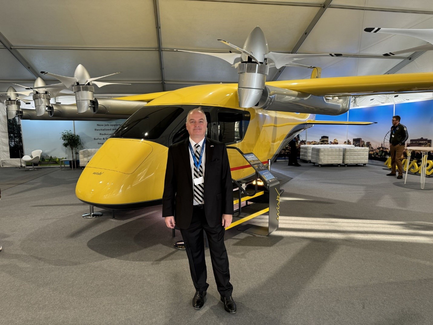 Man in suit standing in front of a yellow electric vertical takeoff aircraft at an expo.