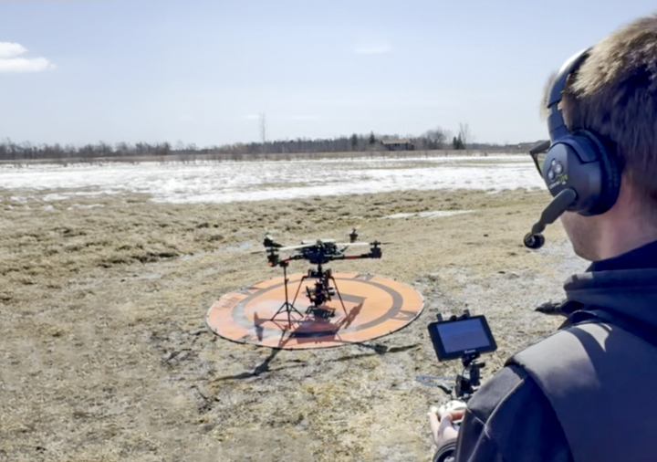 Un opérateur de drone portant un casque et tenant une télécommande se tient dans un champ boueux et partiellement enneigé, se préparant à lancer un drone multirotor reposant sur une piste d'atterrissage circulaire orange et noire.