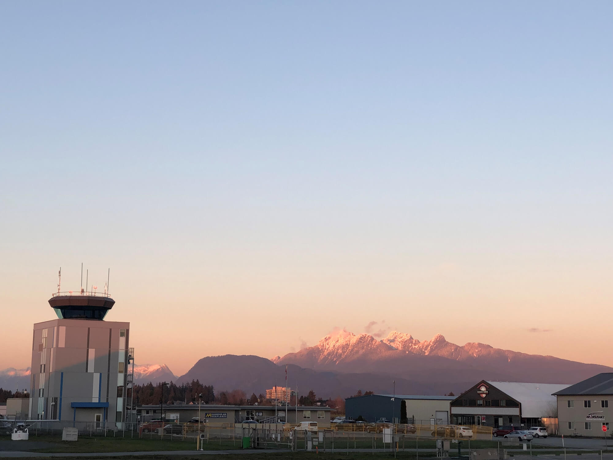 A control tower and several airport buildings are visible at the base of a snow-capped mountain range, all bathed in the warm, pink light of a sunrise.