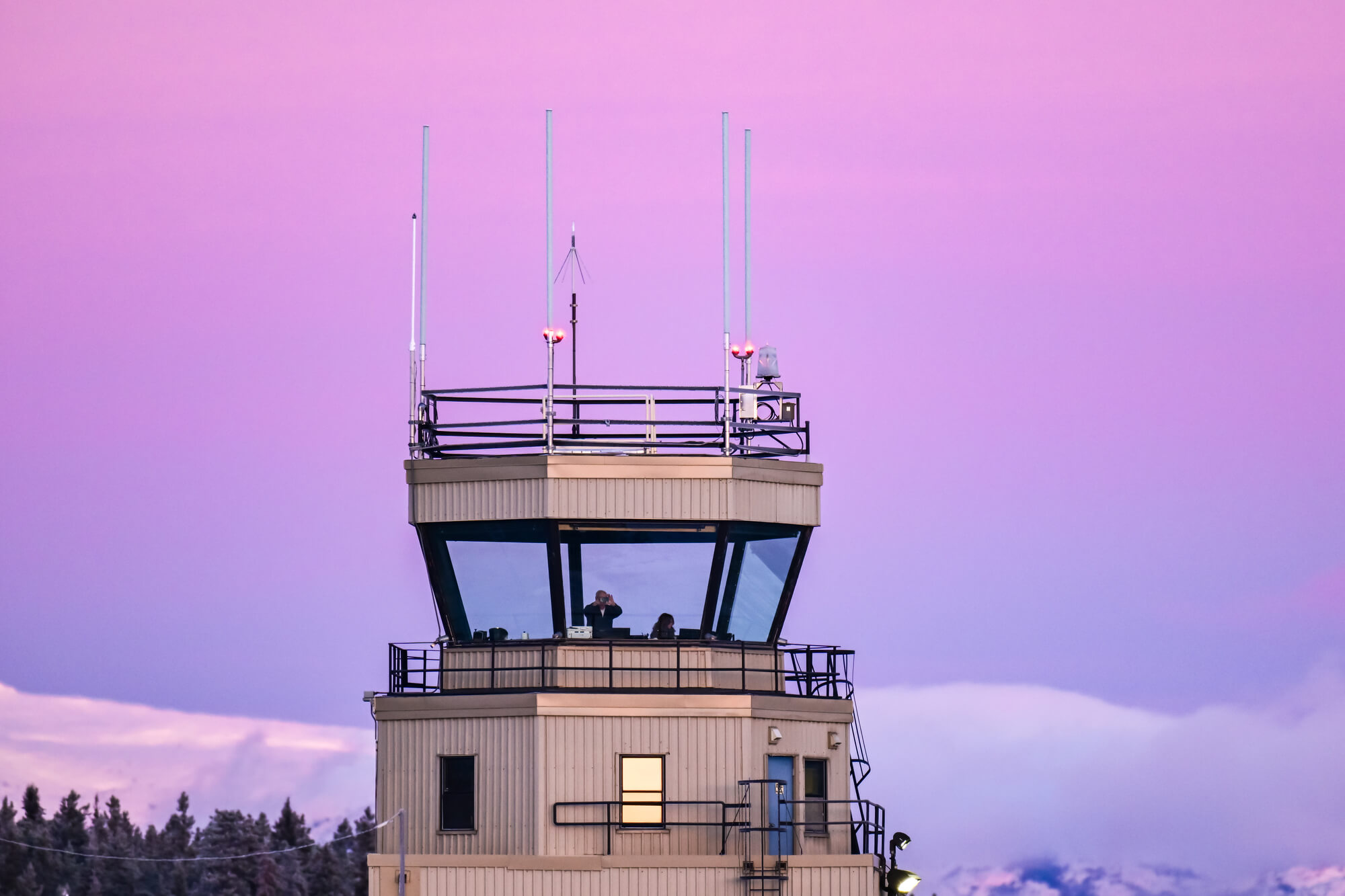 The top of an air traffic control tower shows two people inside the observation deck against a vibrant, purple and pink evening sky.