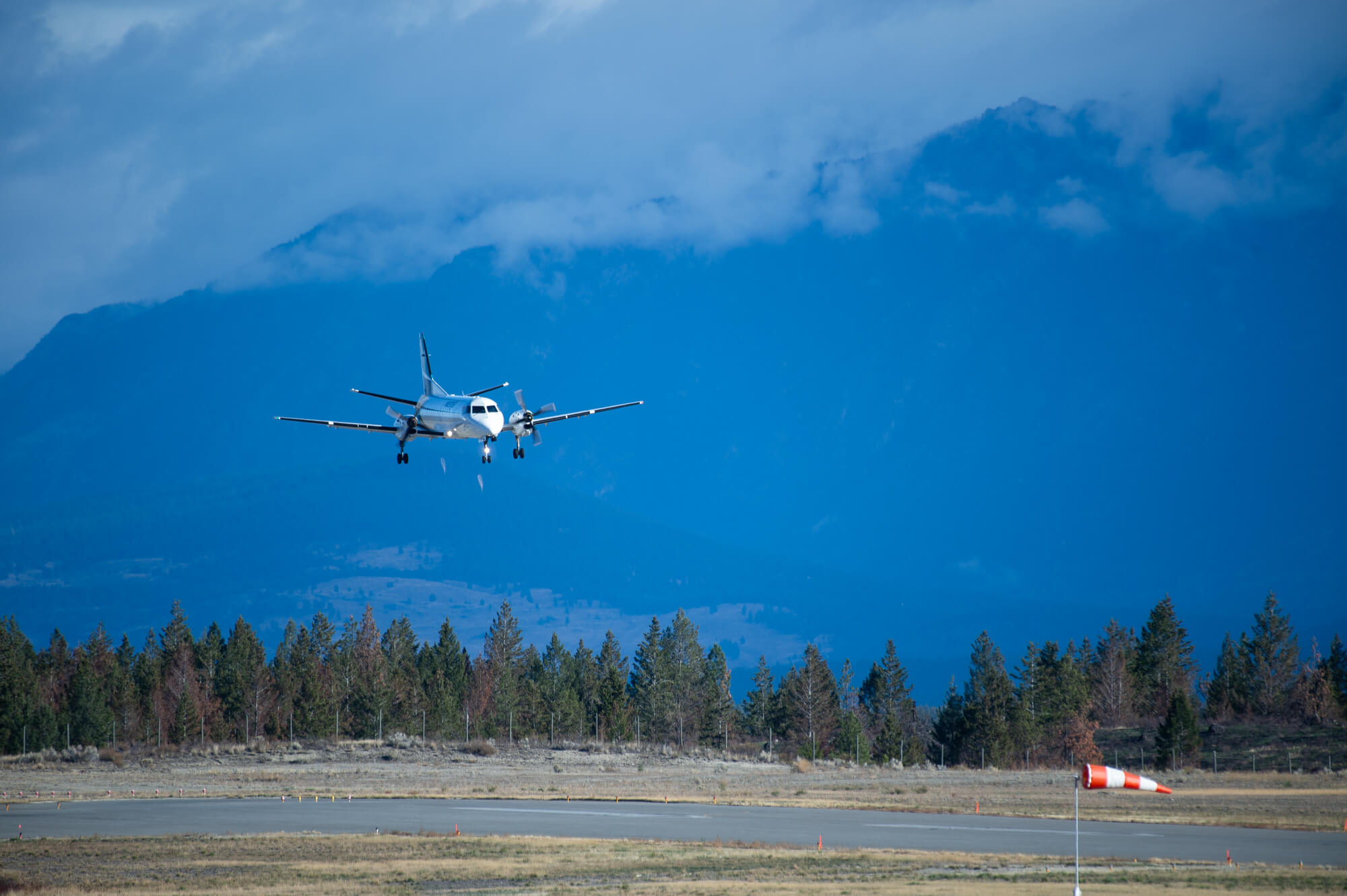 A twin-propeller passenger plane descends toward a runway with a forest in the background and a large, blue mountain shrouded in mist behind it.