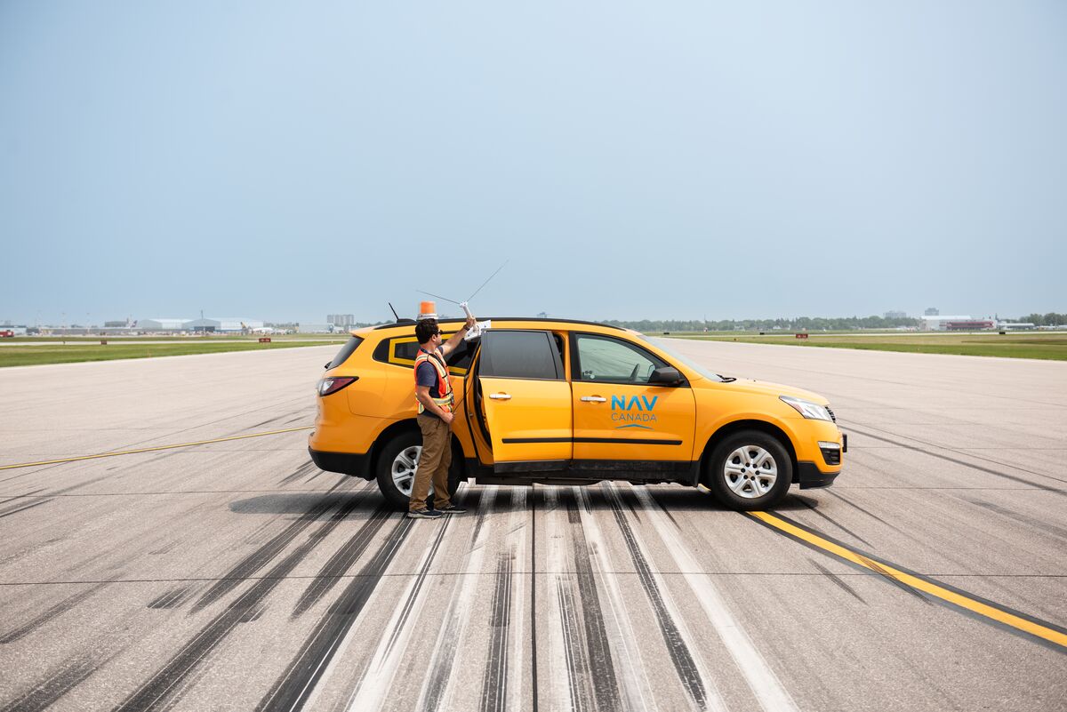 A yellow NAV CANADA vehicle parked on a runway. The vehicle has “NAV CANADA” written on the side. Next to the back door, a person wearing a high-visibility vest is holding an antenna. 