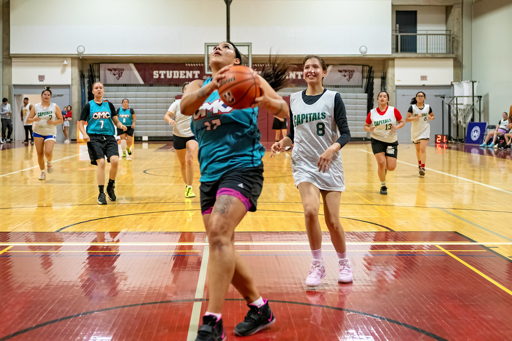 A group of women playing basketball in the gym. At the centre of the photo is a woman aiming the ball towards the net.