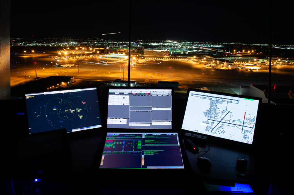 Une tour de contrôle du trafic aérien la nuit avec plusieurs moniteurs affichant des informations de vol et une vue sur une piste d'aéroport bien éclairée.