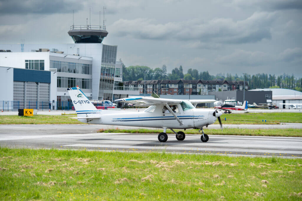 Un petit avion blanc circulant sur une piste d'atterrissage dans un aéroport avec une tour et des hangars en arrière-plan.