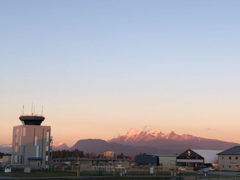 Une tour des services de la circulation aérienne dans un aéroport avec des montagnes enneigées en arrière-plan et un ciel rose au coucher du soleil.