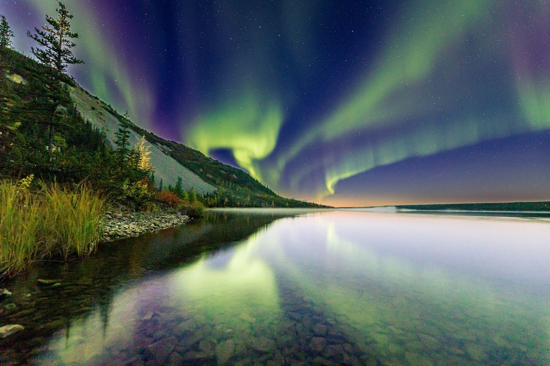 Les aurores boréales brillent dans le ciel et se reflètent sur un lac calme au rivage rocailleux et boisé.