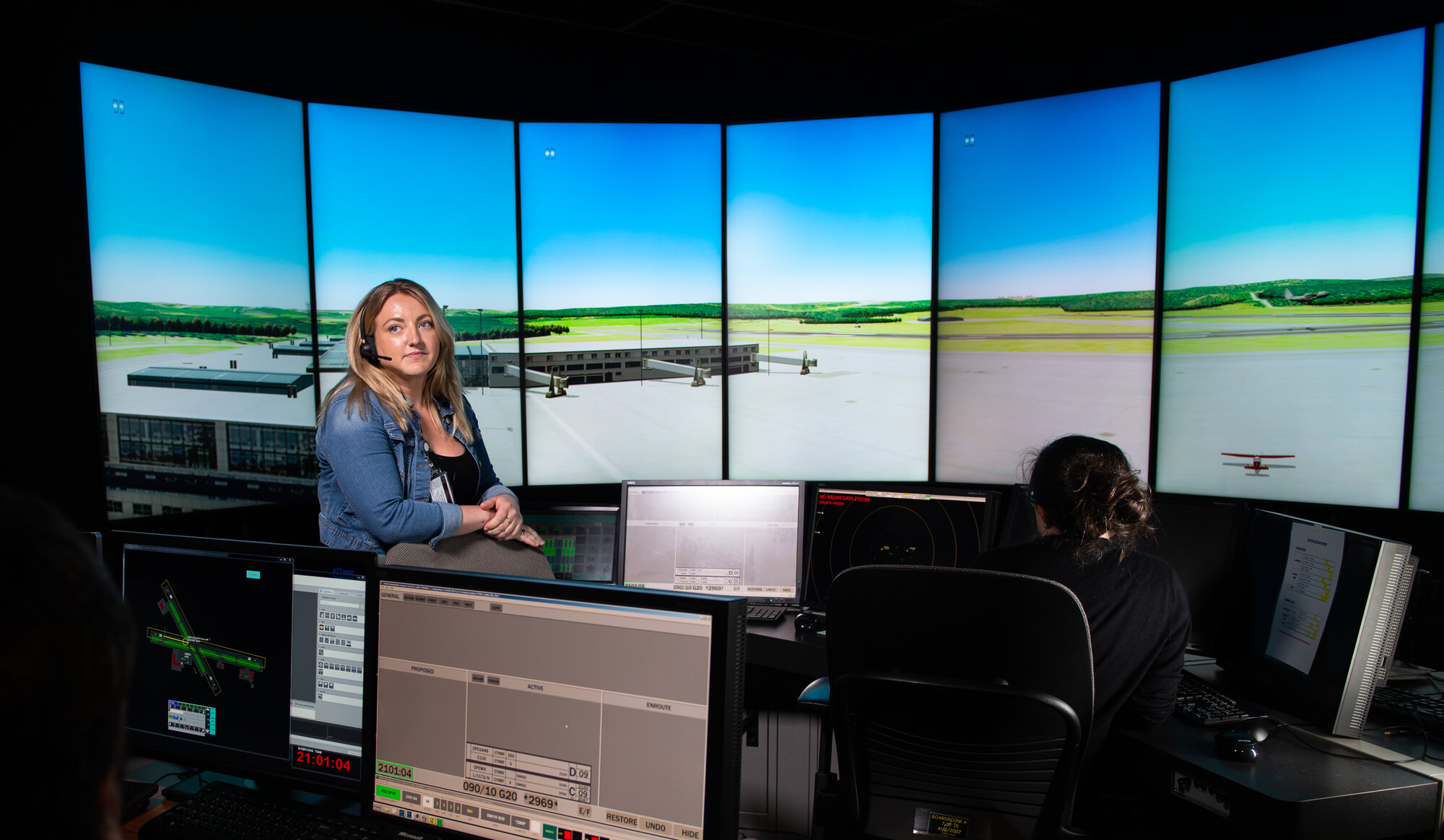 A woman wearing a headset stands in a training room in front of a large, curved screen displaying an airport runway. 