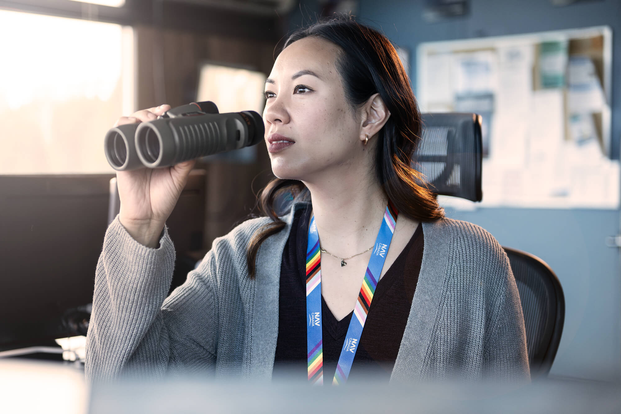 A NAV CANADA employee wearing a lanyard with a rainbow design looks out of a control tower window through binoculars.