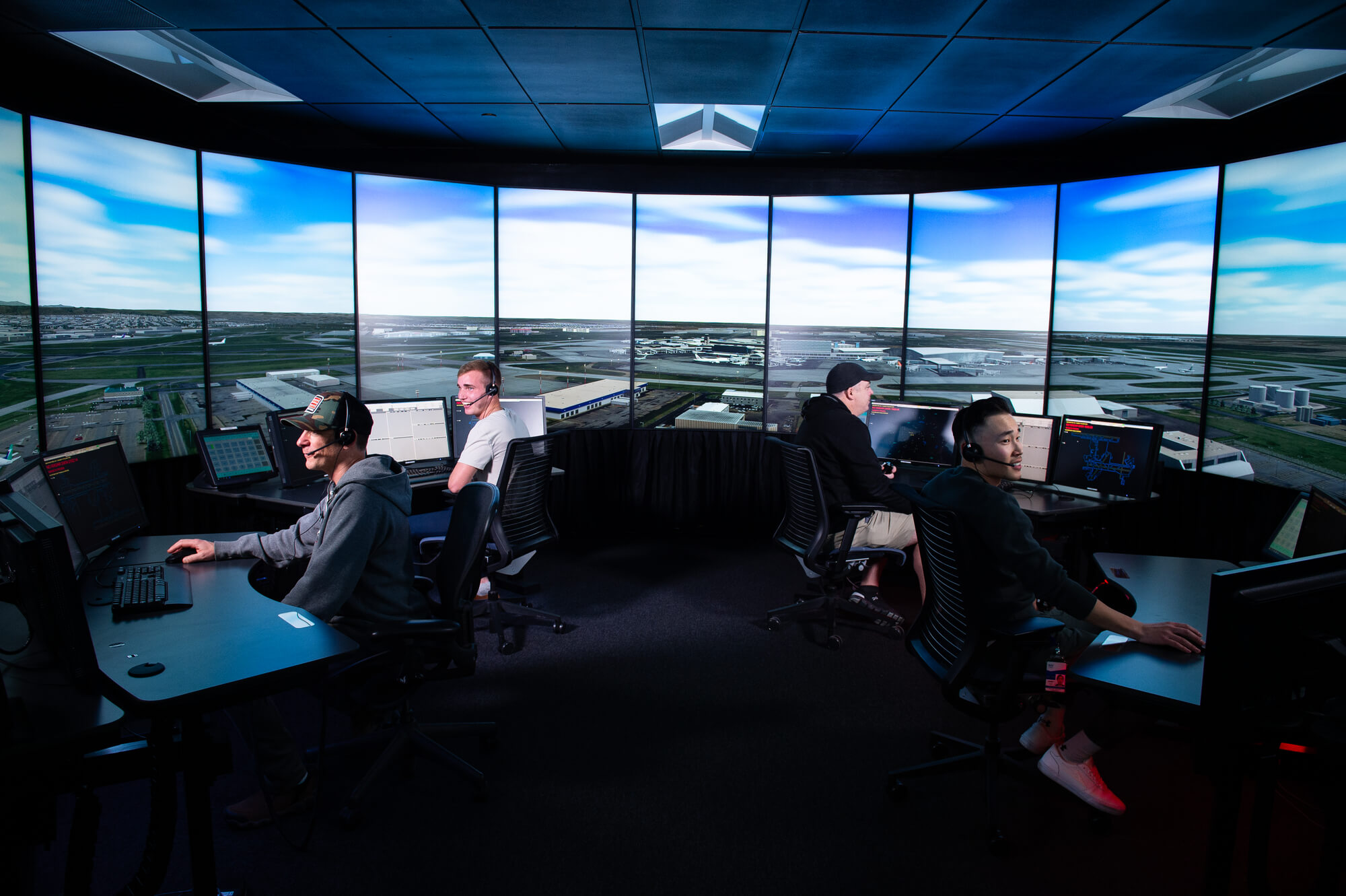 Four individuals wearing headsets operate air traffic control simulators in a training room surrounded by large panoramic screens displaying a virtual airport environment.