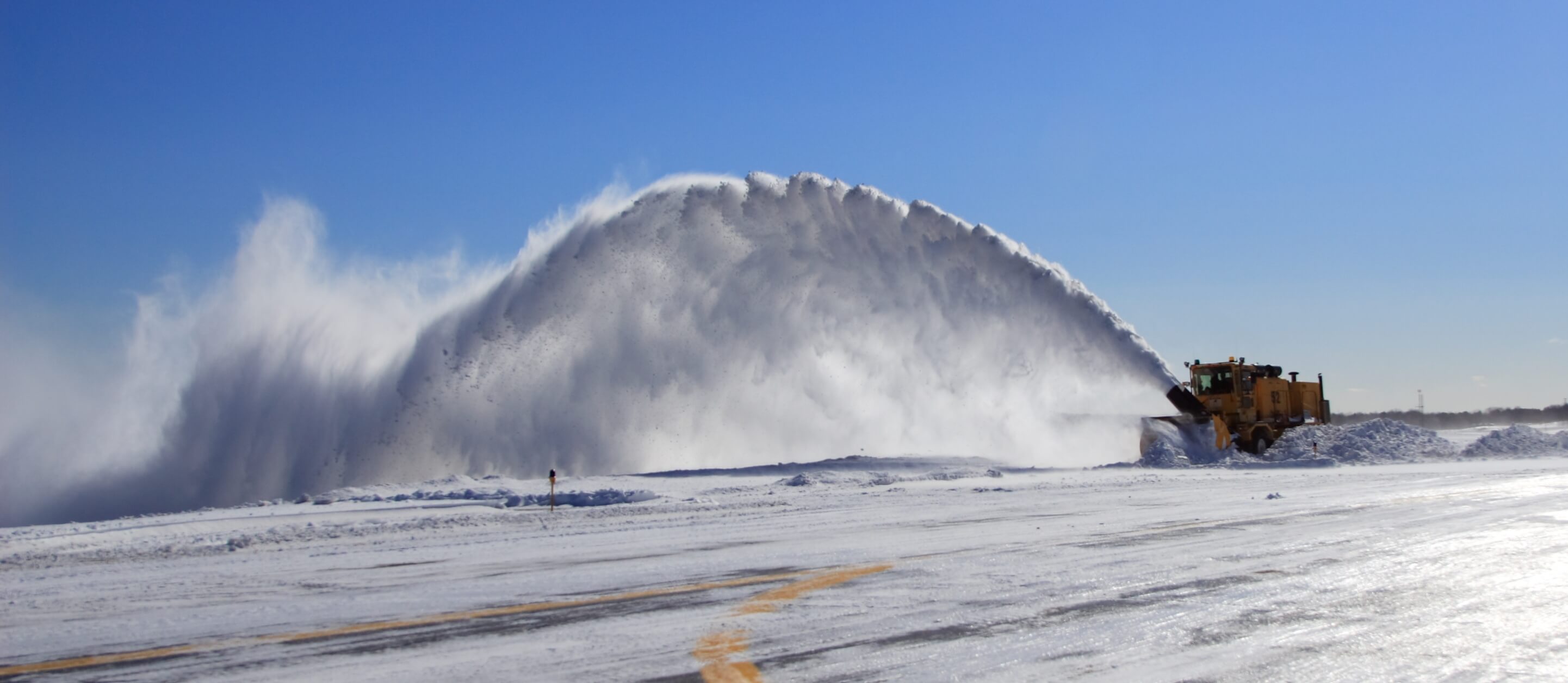 A large yellow snow blower clears a runway at an airport, spraying a high arc of snow into the air.